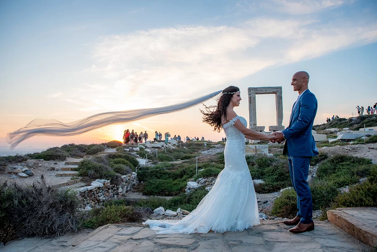 Just married couple holding hands while the woman's veil floats in the air, in front of Portara in Naxos, captured by Naxos Photographer